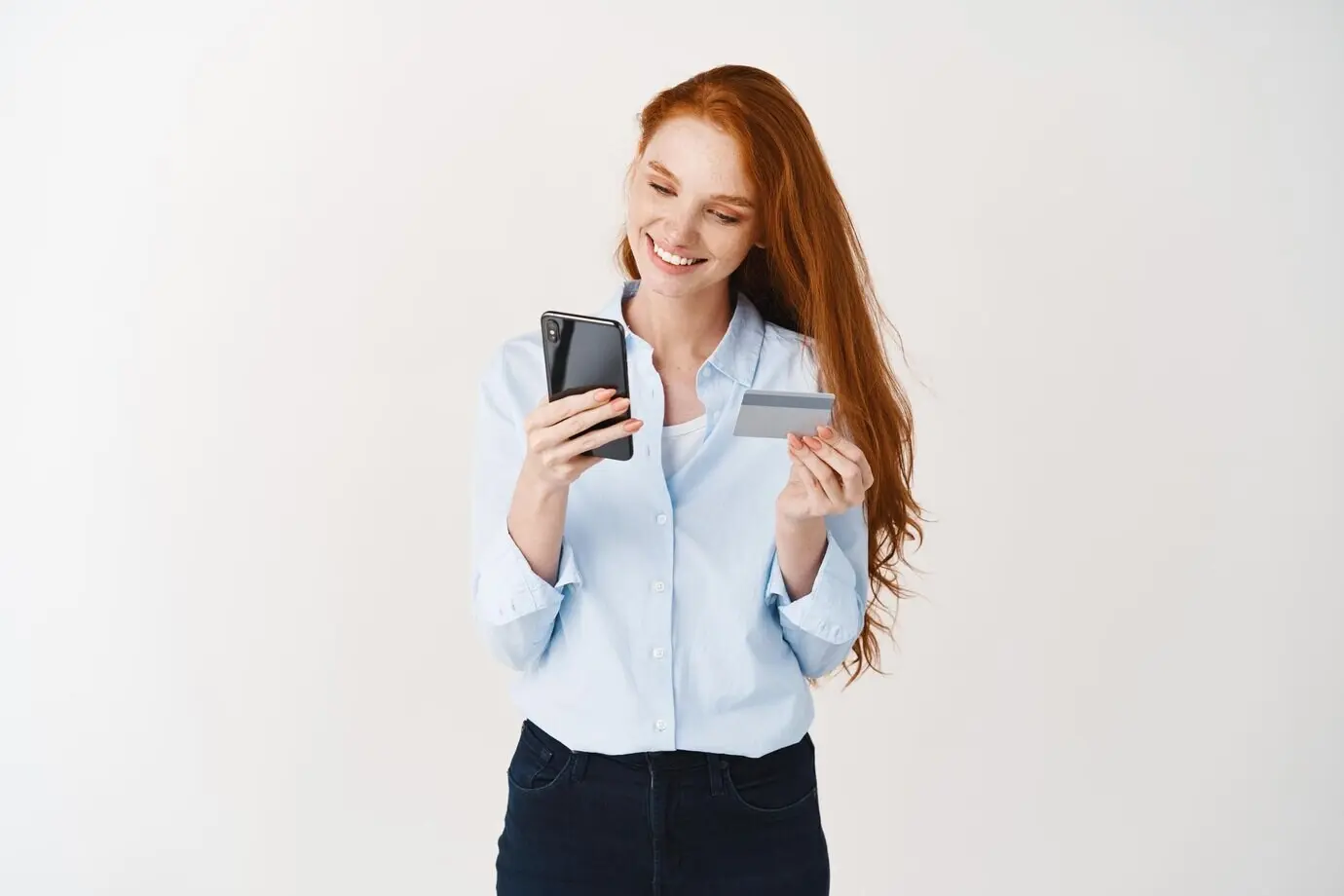 Ein lächelndes Mädchen shoppt online, hält eine Kreditkarte und tätigt einen Kauf per Smartphone vor weißem Hintergrund.