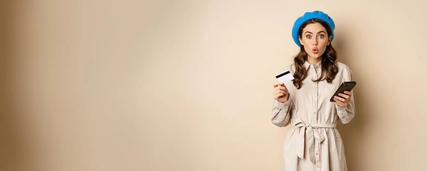 Porträt eines Mädchens, das begeistert schaut, „Wow“ sagt und eine Kreditkarte sowie ein Mobiltelefon hält, vor beigefarbenem Hintergrund stehend.