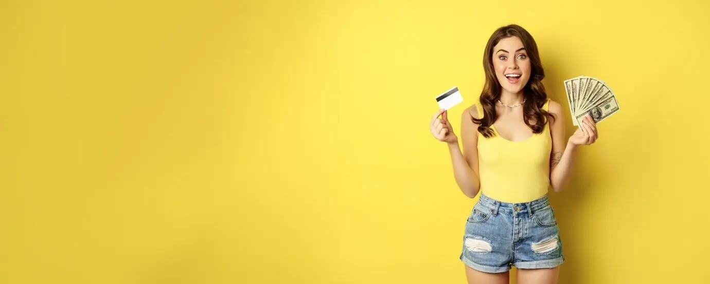 Junge, hübsche Frau in sommerlichem Outfit hält eine Kreditkarte und Bargeld in den Händen und steht vor einem gelben Hintergrund.